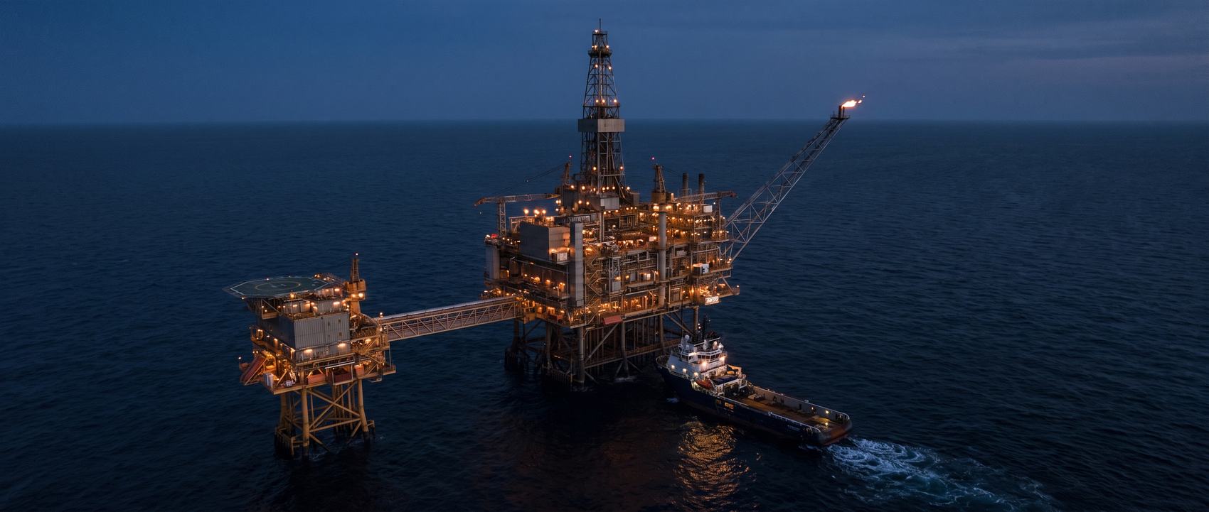 An offshore production platform complex with a drilling derrick and supply vessel at blue hour